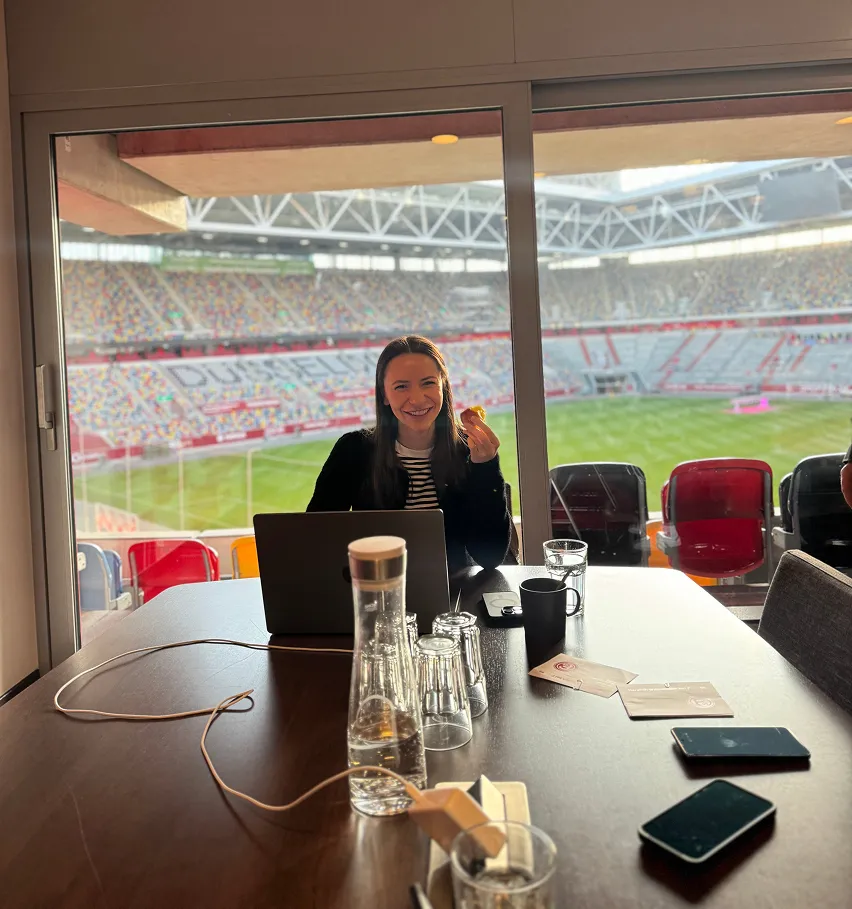 Paula sitzt mit Laptop in einer Stadion-Loge, im Hintergrund das Spielfeld. Sie arbeitet fokussiert und lächelt in die Kamera.