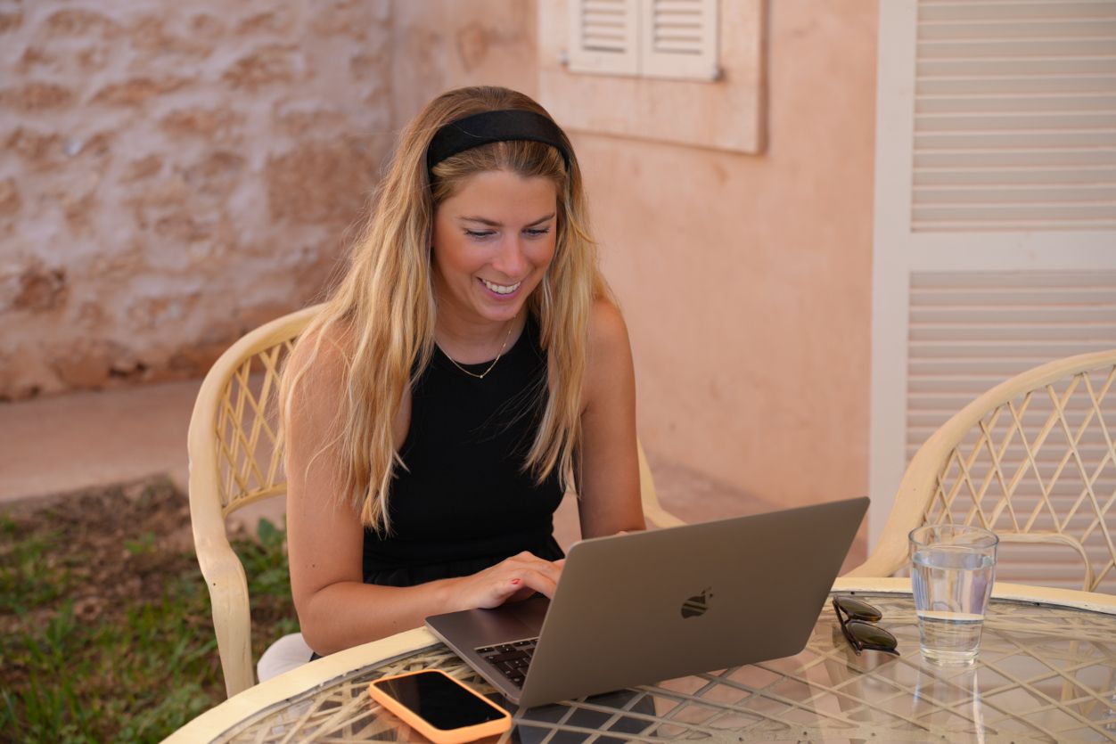 Theresa sitzt mit Laptop im Freien, fokussiert auf den Bildschirm. Ein sonniger Arbeitstag in mediterraner Umgebung bei der Workation.